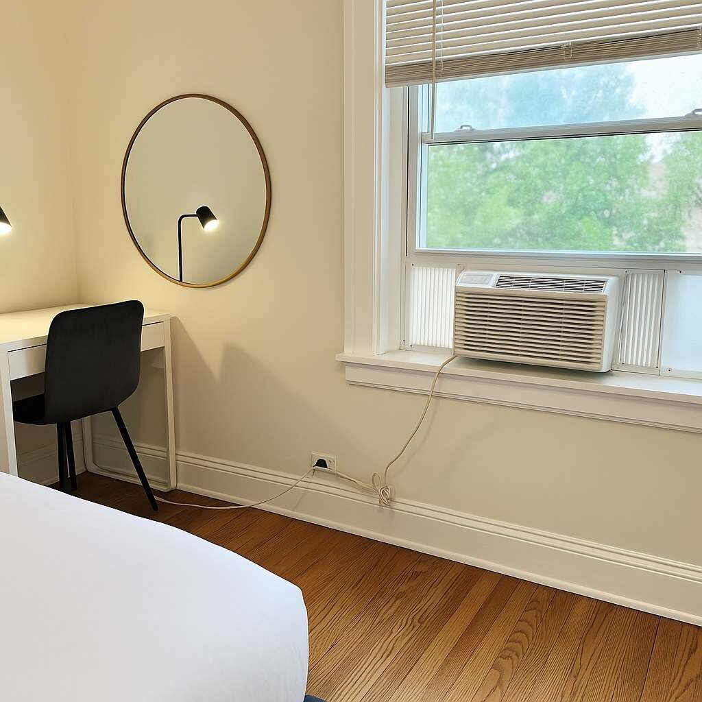A bedroom with a white bed, a black chair, a round mirror, a window with blinds, and an air conditioner.