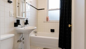 Ein Badezimmer mit weißem Waschbecken, WC und Badewanne. Ein Fenster bietet Blick ins Freie, und eine kleine Ablagefläche zeigt Toilettenartikel.
