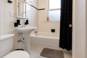 A bathroom with a white sink, toilet, and bathtub. There is a window with a view of the outside, and a small shelf with toiletries. - Ideal & Spacious 1BR Apt (Chicago)