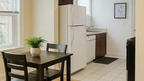 Eine kleine Küche mit Kühlschrank, Spüle und Schränken. Im Vordergrund befindet sich ein Esstisch mit Stühlen und ein Teppich.