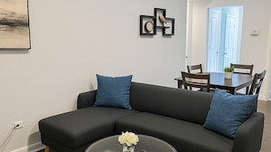 Een moderne woonkamer met een hoekbank, een glazen salontafel en een eethoek met een tafel en stoelen.