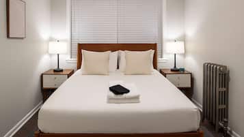 A neatly made bed with a wooden headboard, two bedside tables with lamps, a radiator, and a framed picture on the wall.