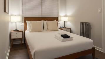 A neatly made bed with a wooden headboard, two bedside tables with lamps, a radiator, and a window with blinds.