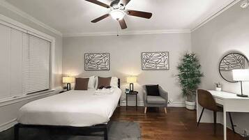 Habitación de hotel con cama, escritorio, silla, ventilador de techo y dos cuadros en la pared.