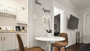 Een moderne keuken met witte kasten, een witte ronde tafel en twee bruine stoelen. Er staat een witte orchidee op de tafel en op de muur staat een bordje met de tekst 'Bon Appetit'.
