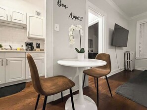 Ett modernt kök med vita skåp, ett vitt runt bord och två bruna stolar. På bordet står en vit orkidé och på väggen hänger en skylt med texten ”Smaklig måltid”.