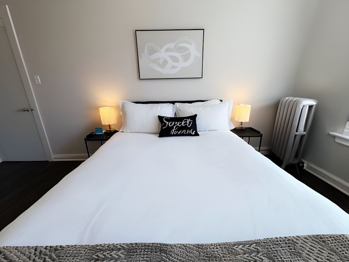 Cama bem arrumada com roupa de cama branca, duas luminárias de cabeceira, uma obra de arte abstrata emoldurada acima da cama e um radiador à direita.