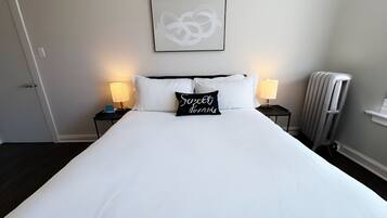 Cama bem arrumada com roupa de cama branca, duas luminárias de cabeceira, uma obra de arte abstrata emoldurada acima da cama e um radiador à direita.