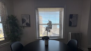 Een ronde tafel met twee stoelen, een potplant en een vaas met bloemen. Twee ramen met zonwering, een radiator, en twee ingelijste kunstwerken aan de muren.