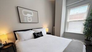 Een hotelkamer met een bed, nachtlampjes, een kamerplant en een raam met jaloezieën.