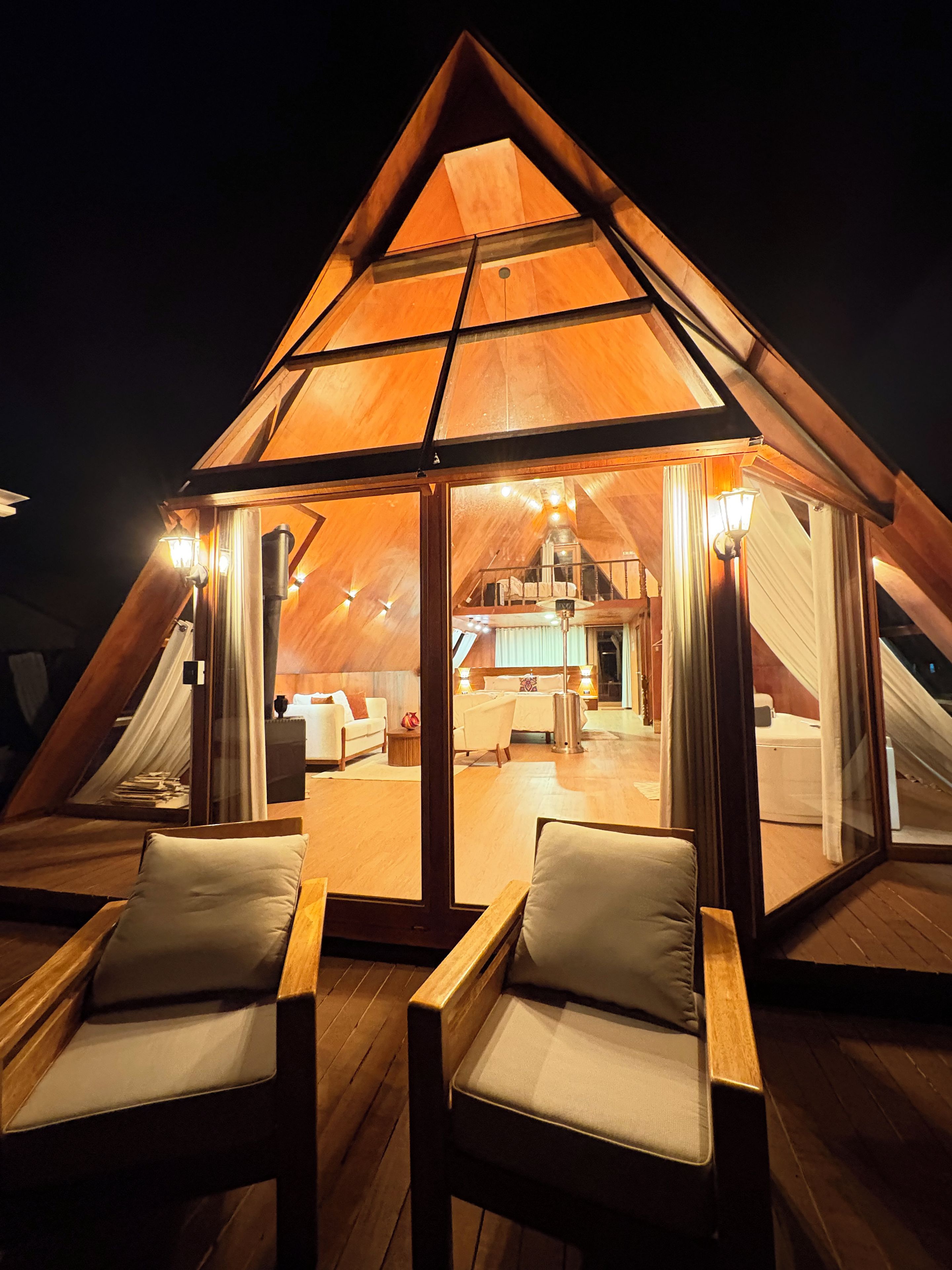 Una estructura triangular de madera con cerramiento de vidrio, amoblada con una cama, un sofá y una mesa de comedor.