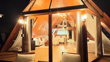 Una estructura triangular de madera con cerramiento de vidrio, amoblada con una cama, un sofá y una mesa de comedor.