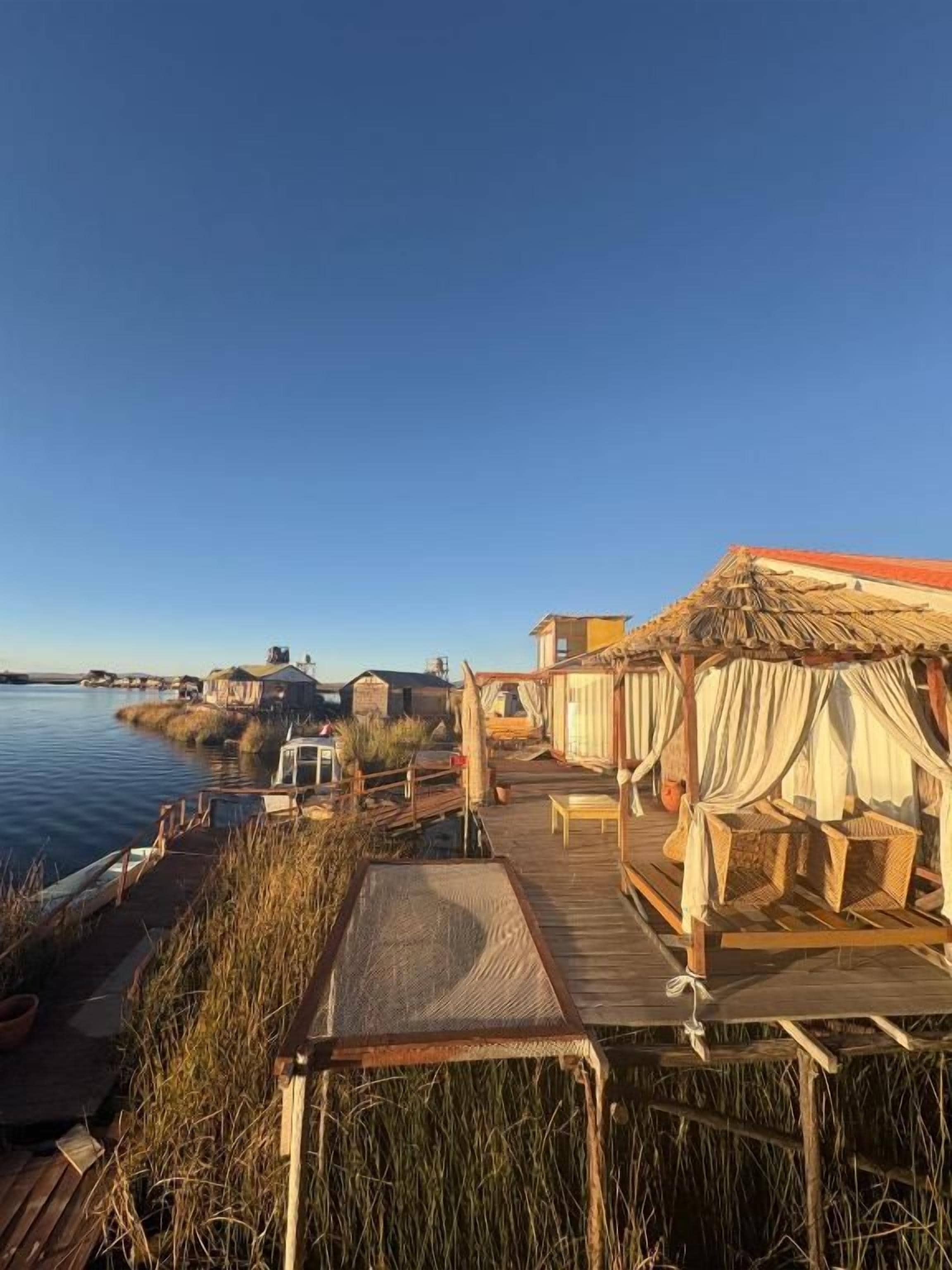 Una terraza de madera con mobiliario de mimbre, con vistas a un cuerpo de agua tranquilo y una costa lejana.