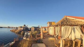 Una terraza de madera con mobiliario de mimbre, con vistas a un cuerpo de agua tranquilo y una costa lejana.