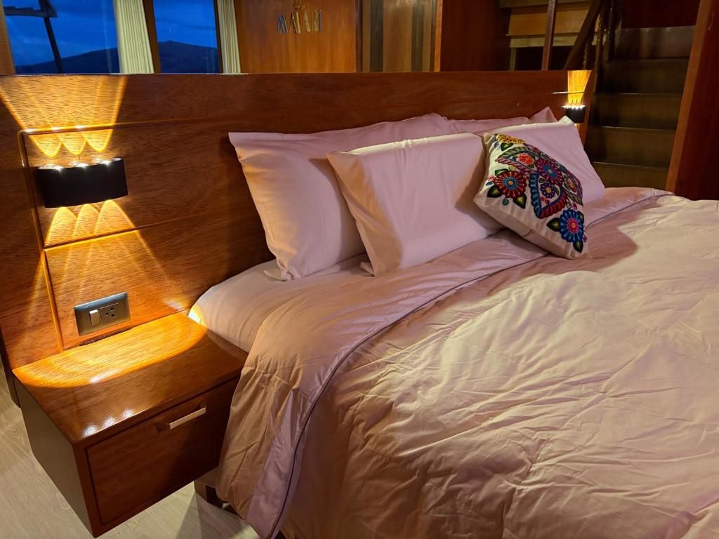 Habitación de hotel con cabecera de madera, mesita de noche con lámpara, cama con ropa blanca y almohada colorida.