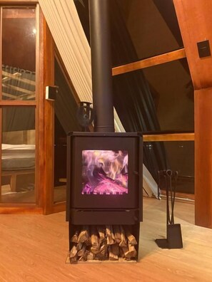 A wood-burning stove with a visible fire, placed in a room with a sloped ceiling and wooden beams.