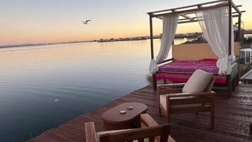 A wooden deck with a bed and chairs overlooking calm water.