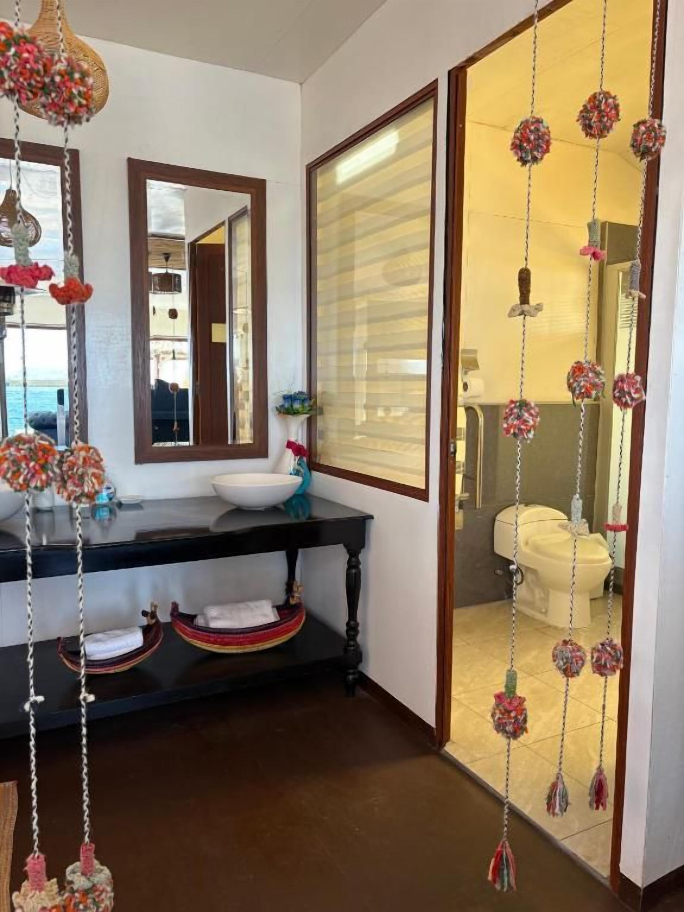 A bathroom with a black vanity, a white sink, and a mirror. There are decorative hanging items and a window with blinds.