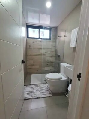 A small bathroom with a toilet, a shower, and a window. - Stylish Metropolitan 2 Bedroom Apartment (Santo Domingo)