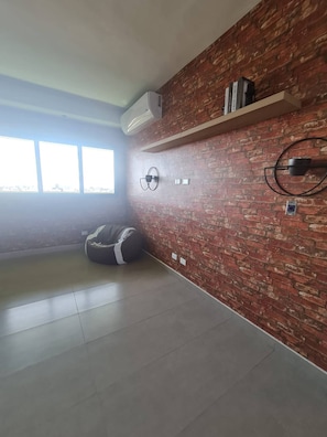 A room with a brick wall, a bean bag chair, a shelf with books, and a window. - Stylish Metropolitan 2 Bedroom Apartment (Santo Domingo)