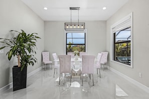 A dining room with a white table and chairs, a large window with a view, and a hanging light fixture. - Villa du Soleil | Luxury, Pool & Palm Dreams (Cape Coral)