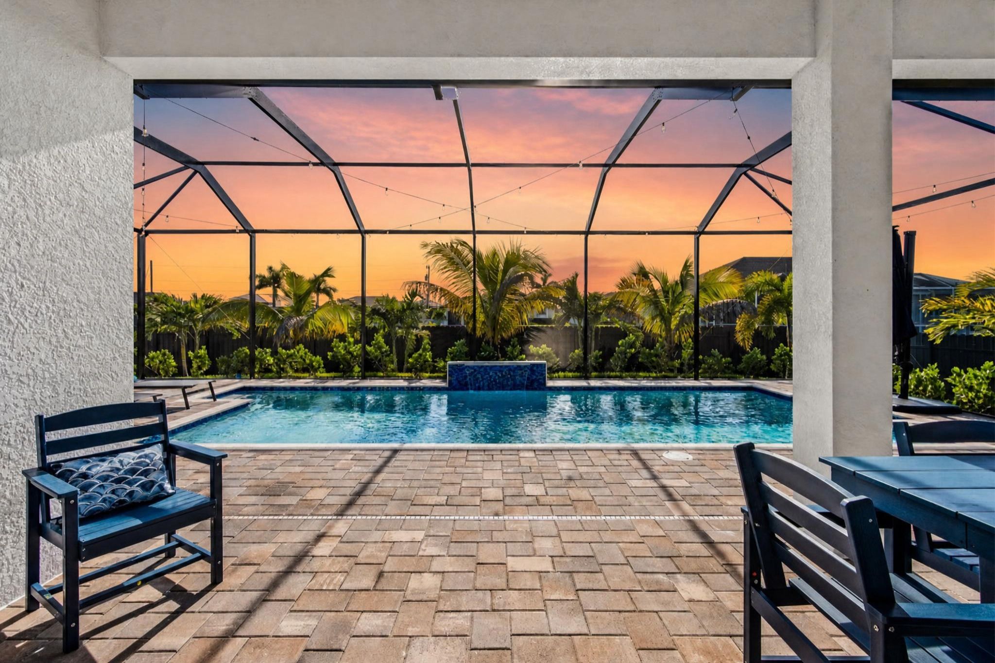 An outdoor swimming pool with a brick patio, surrounded by palm trees and a sunset view.