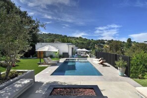 A rectangular swimming pool with lounge chairs and umbrellas, surrounded by a landscaped garden and a view of a hillside. - Round Valley Residence (Sherman Oaks)