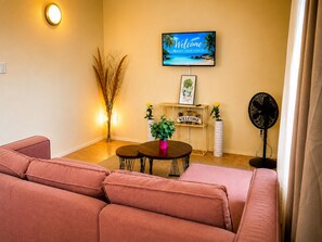 Ein Wohnzimmer mit einer Couch, einem kleinen Tisch mit Pflanze, einem Willkommensschild, einem Fernseher der eine Strandansicht zeigt und einem Standventilator.