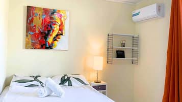 A hotel room with a bed, a painting, an air conditioning unit, a bedside table with a lamp, and a shelf with toiletries.
