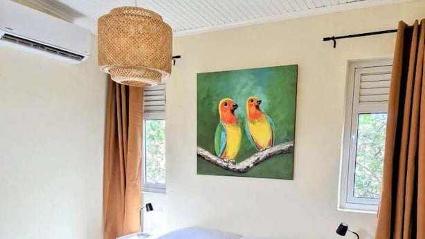 A bedroom with a bed, a nightstand, a lamp, a painting of birds, and a window with curtains. - Apartment Near Beaches, Dining & Attractions (Willemstad)