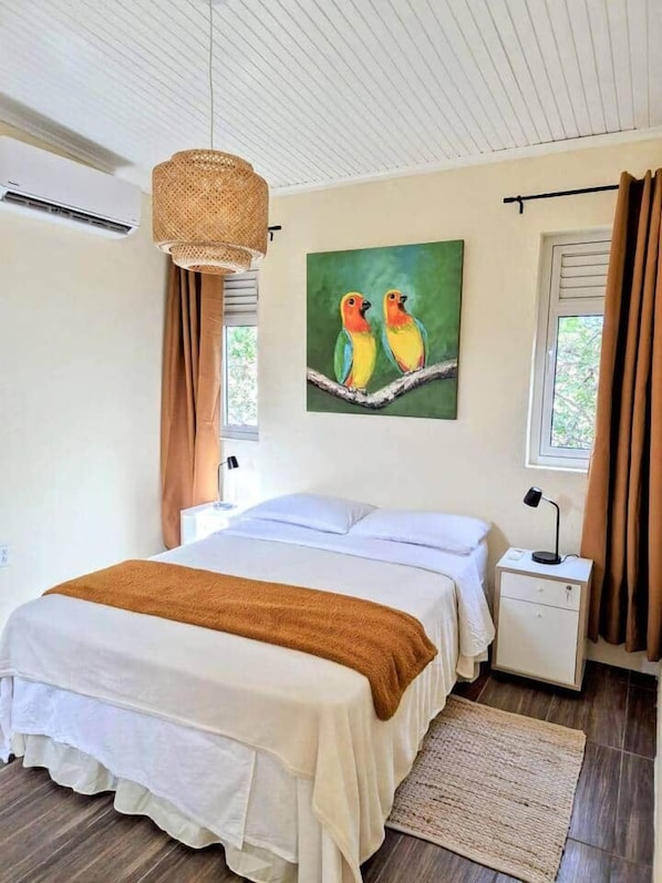 Ein Schlafzimmer mit einem Bett, einem Nachttisch, einer Lampe, einem Vogelbild und einem Fenster mit Vorhängen.