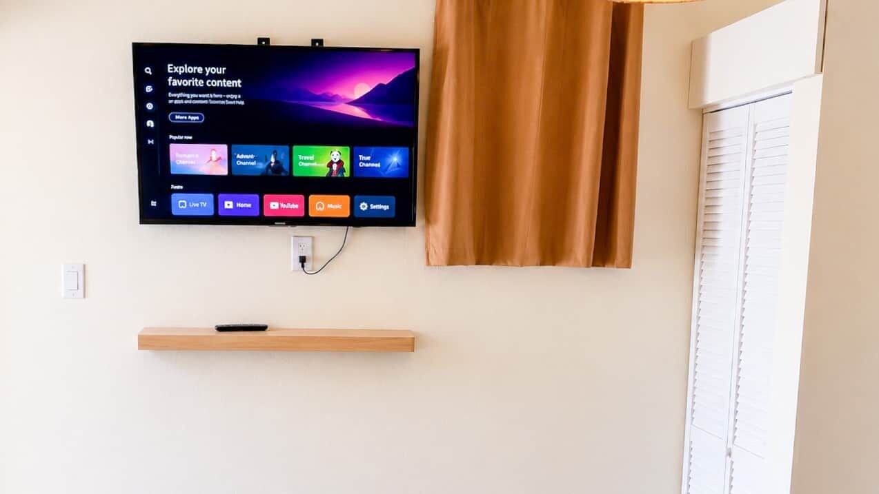 Een flatscreen televisie bevestigd aan een muur, met daaronder een houten plank, een raam met zonwering en een gordijn aan de rechterkant.