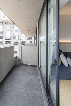 A balcony with a view of a building complex, a glass door leading to a room with a sofa and cushions, and a tiled floor. - Girasol - Loft Condesa - 01B (Mexico City)