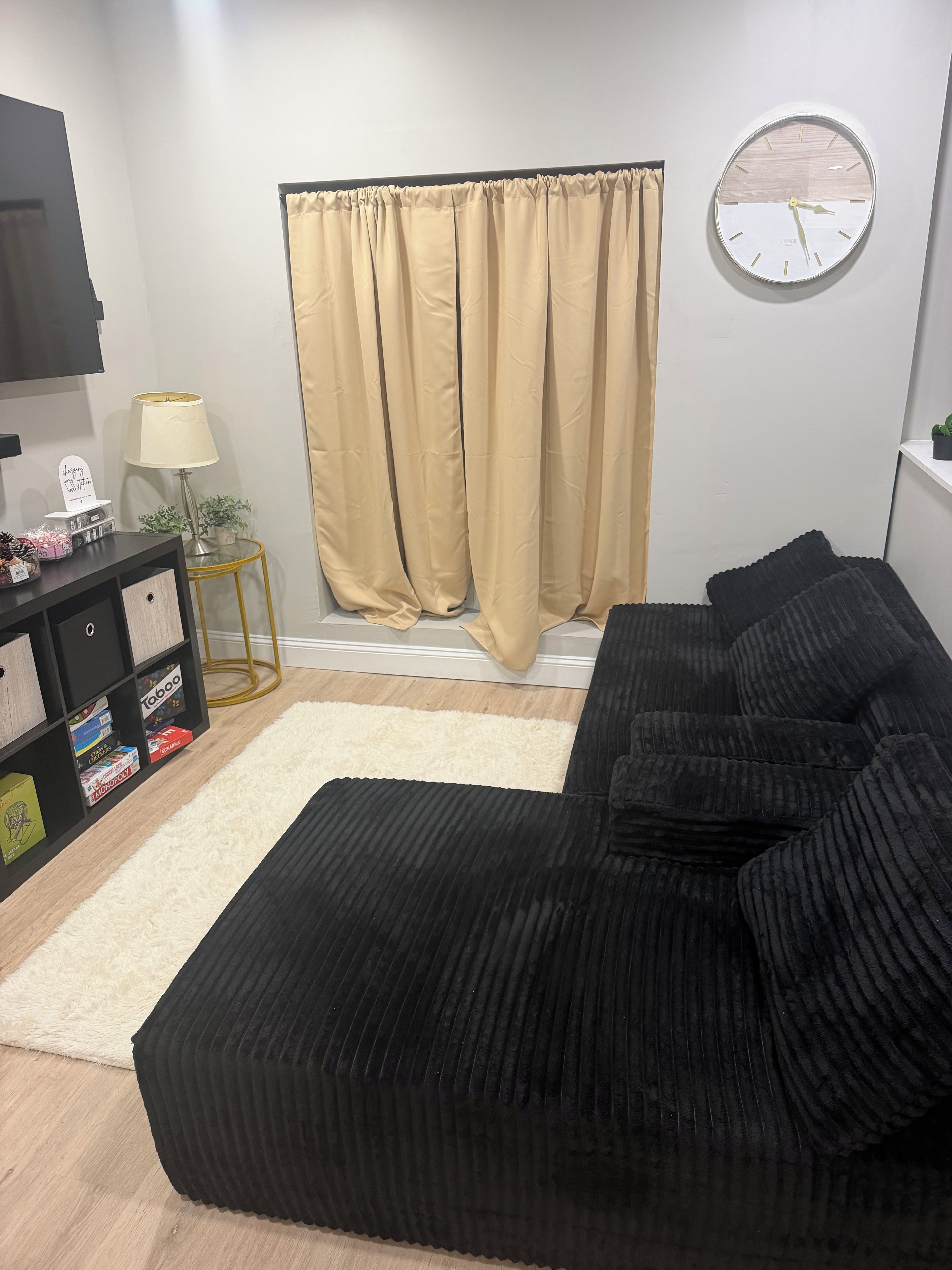 Een moderne woonkamer met een zwarte bank, een ronde wandklok, een beige gordijn, een klein bijzettafeltje met een lamp en een zwarte entertainmentkast met diverse spullen.