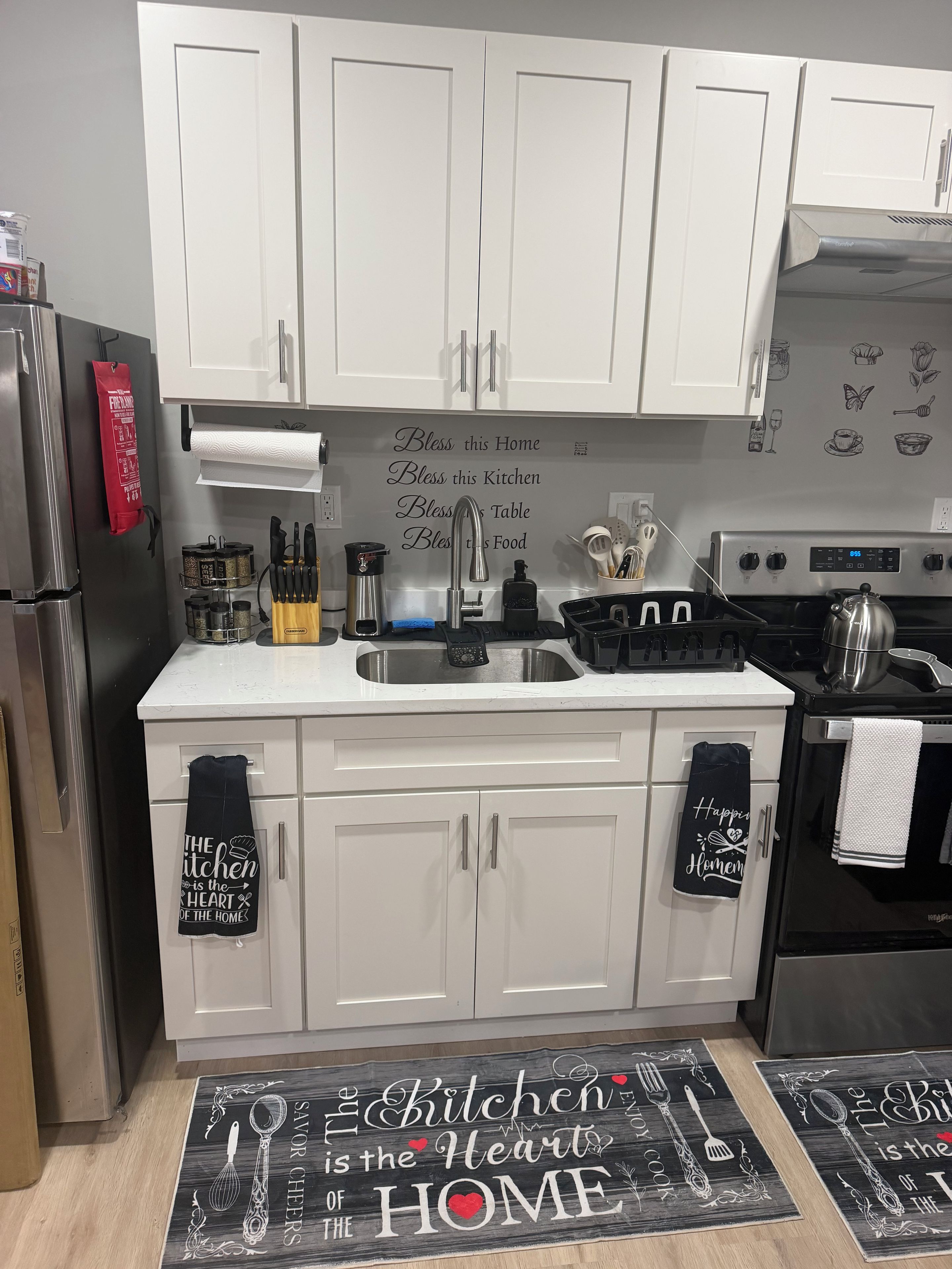 A kitchen with white cabinets, stainless steel appliances, and kitchenware. There are two mats on the floor with kitchen-related text.