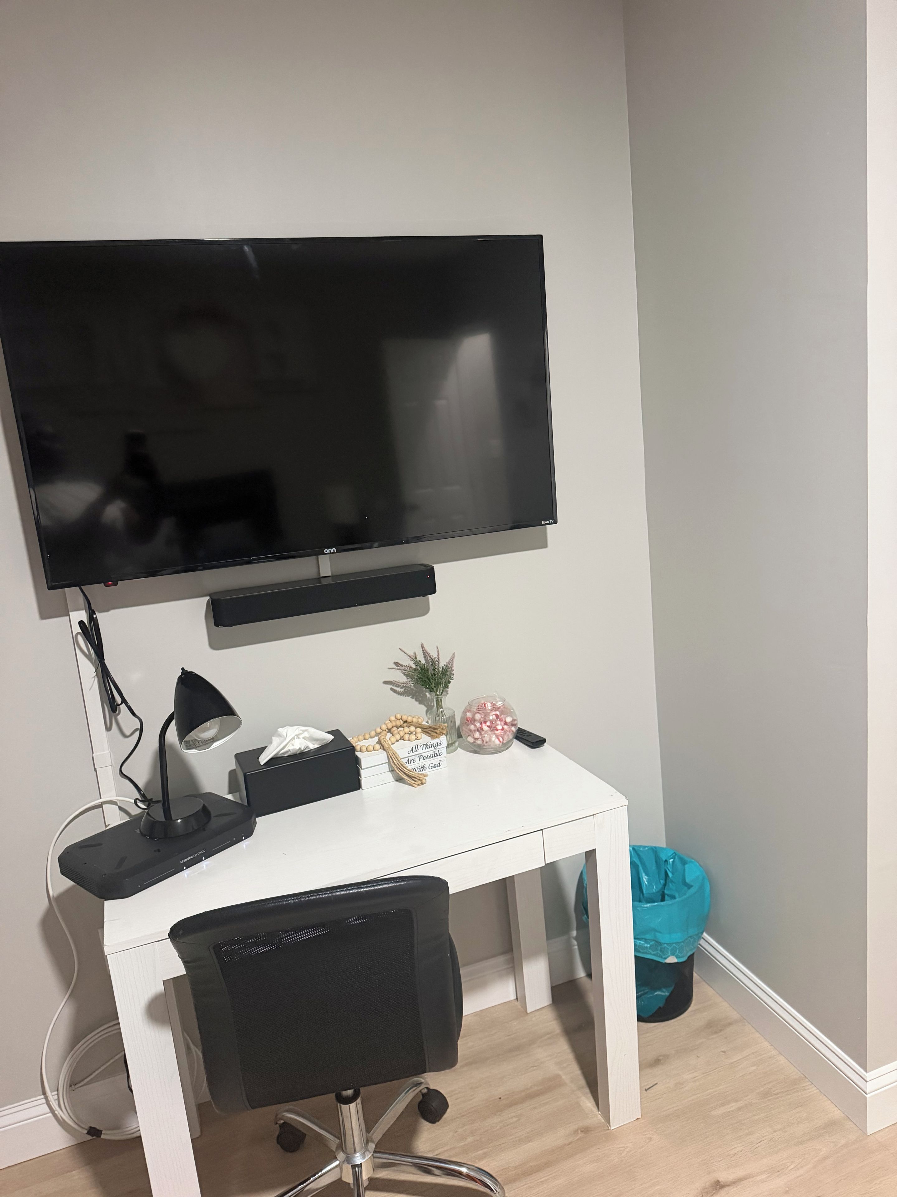 A white desk with a black chair, a flat-screen TV mounted above, a desk lamp, a small vase with a plant, and a decorative item with a message.