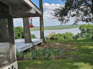 Une terrasse en bois avec une mangeoire à oiseaux, une vue sur un lac et, au loin, un cerf qui broute.