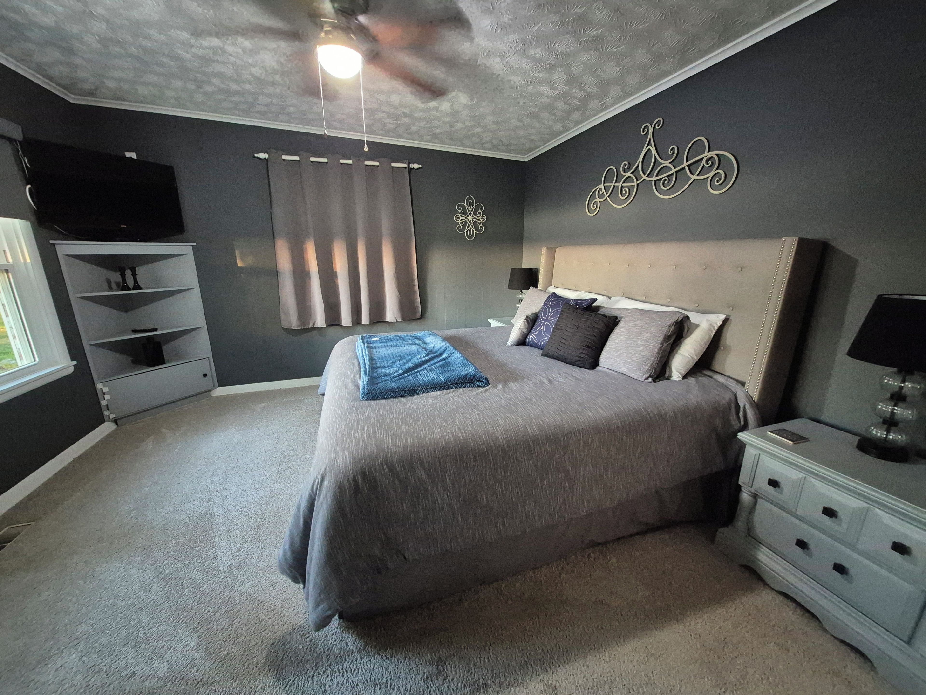 A bedroom with a large bed, a ceiling fan, a TV mounted on the wall, and a nightstand with a lamp.