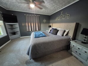 A bedroom with a large bed, a ceiling fan, a TV mounted on the wall, and a nightstand with a lamp.