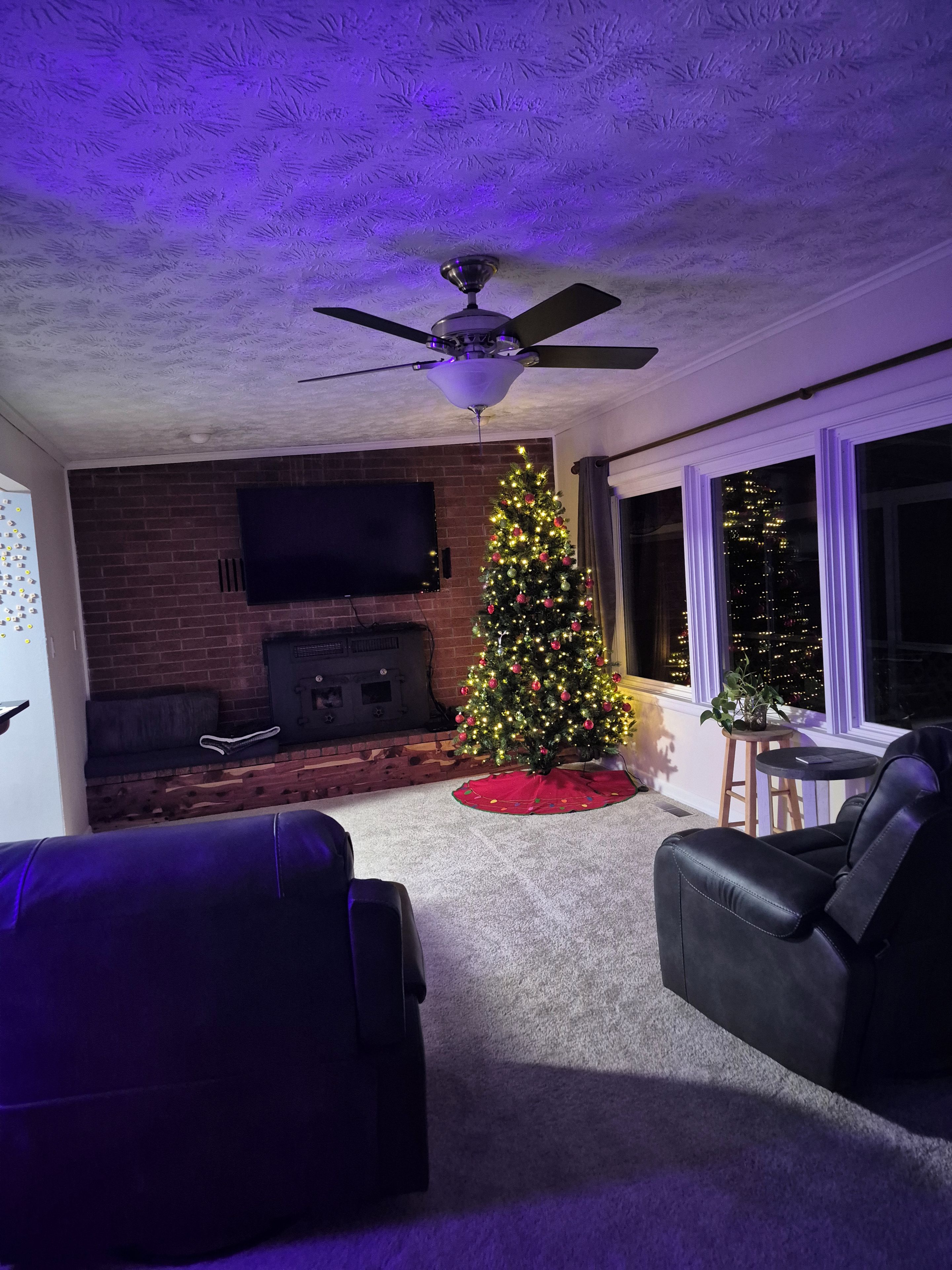 A living room with a fireplace, a Christmas tree, a ceiling fan, and a large window.