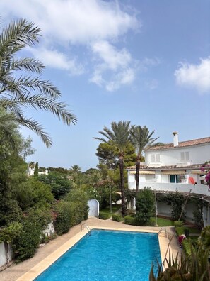 A swimming pool with a clear blue water, surrounded by lush greenery and a white building with a tiled roof. - GuestReady - Coastal Apartment in Dénia (Denia)