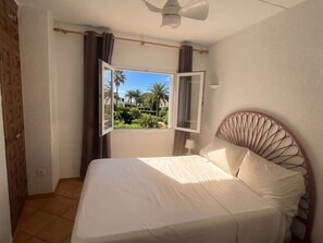 A bedroom with a bed, a window with curtains, a ceiling fan, and a view of greenery outside. - GuestReady - Coastal Apartment in Dénia (Denia)
