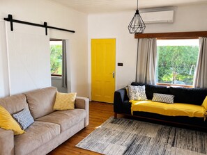 A room with a beige sofa, a black sofa with yellow cushions, a wooden floor, a yellow door, and a window with curtains.