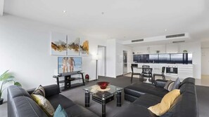 A modern living room with a sofa, a coffee table, and a TV. There is a kitchen in the background with white cabinets and a view of the ocean. - Circle On Cavil Apartment 2146 (Surfers Paradise)