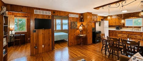 Ein geräumiger Wohnbereich mit Holzboden, einem Fernseher, einem Esstisch mit Stühlen und einer Küchenzeile mit Holdekor und einem Kühlschrank.