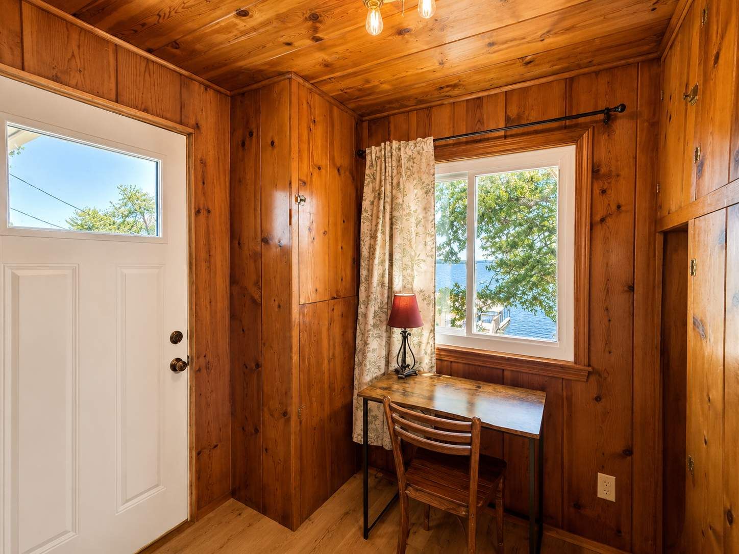 Un interior de madera con una puerta blanca, una ventana que ofrece vistas al exterior, una mesa con una silla y una lámpara.