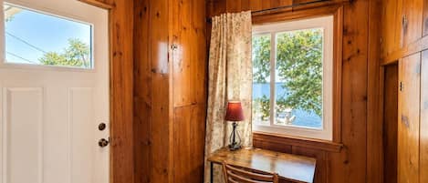 Un interior de madera con una puerta blanca, una ventana que ofrece vistas al exterior, una mesa con una silla y una lámpara.