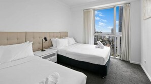 A hotel room with two beds, a nightstand, a lamp, and a view of a cityscape through large windows. - Chevron Renaissance Apartment 1262 (Surfers Paradise)