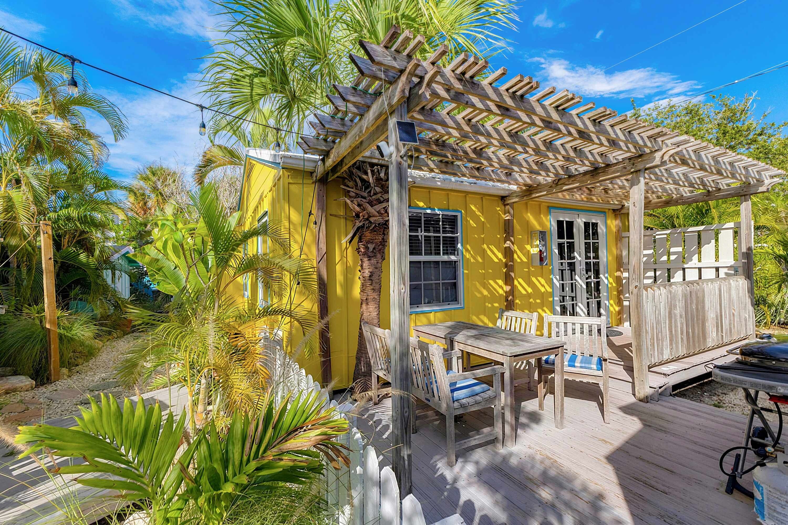 Una piccola casa gialla con un portico in legno, circondata da piante tropicali.