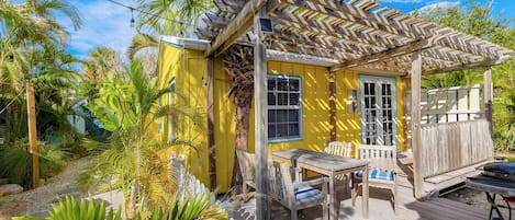 A small yellow house with a wooden porch, surrounded by tropical plants.